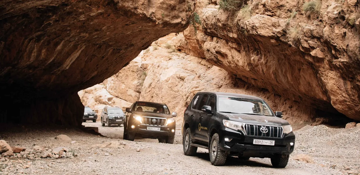 Xped Global: a convoy of branded expedition vehicles passing beneath a canyon arch.