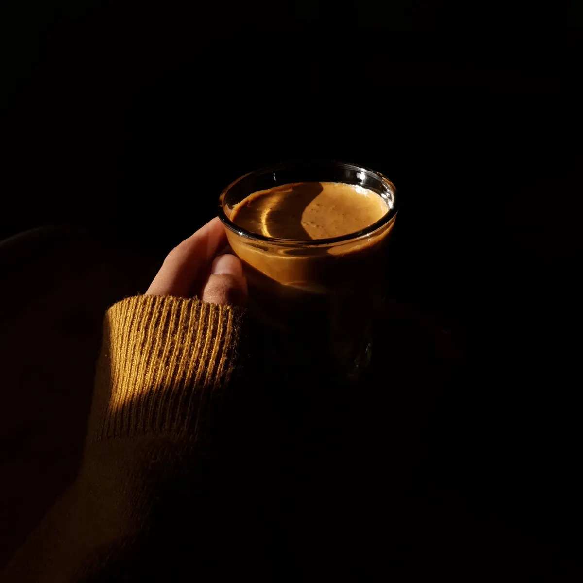 A hand in a mustard jumper holding a small glass of espresso, lit by a single shaft of light in an otherwise dark room.